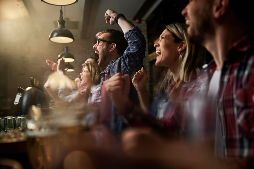 Cheerful fans having fun while screaming and celebrating the success of their sports team while watching a game on TV in a bar. Focus is on man with eyeglasses.