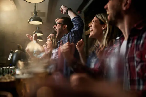 Cheerful fans having fun while screaming and celebrating the success of their sports team while watching a game on TV in a bar. Focus is on man with eyeglasses.