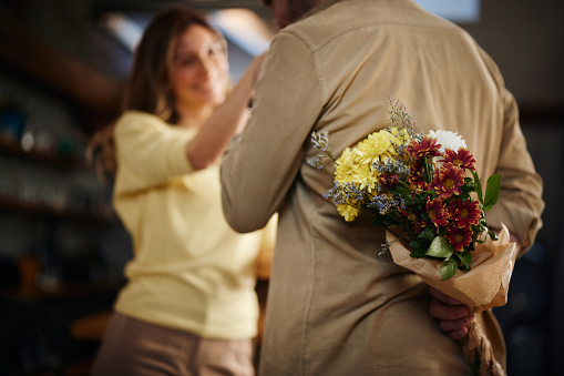 Back view of unrecognizable man hiding flower bouquet behind his back for his wife at home.