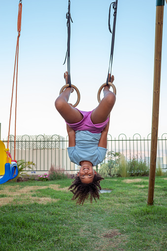 A multiracial curly little girl flipping upside down on gymnastic rings. The smiling elementary-age child wearing a blue t-shirt and shorts, exercising, putting her legs in the gym rings in the back yard of her house. Happy childhood, active lifestyle concept.