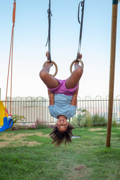 zabawna szczęśliwa mała kręcona dziewczyna huśta się do góry nogami na zewnątrz, trzymając i zwisając z pierścieni gimnastycznych, wkładając w nie nogi. - child swinging playground african descent zdjęcia i obrazy z banku zdjęć
