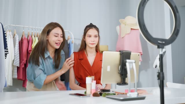 Asian two women vlogger talk to camera to review the cosmetic at home. Beautiful Beauty blogger Influencer woman friends live streaming and showing makeup brush to followers on tablet in office store.