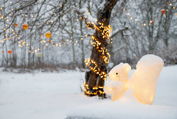 Acrylic artificial squirrel figurine illuminated as Christmas decoration outdoors in home garden, led party lights wrapped around apple tree in snowy winter.