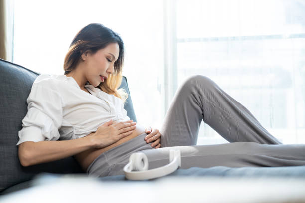 asian young beautiful pregnant woman applying headphones on her belly. attractive expectant pregnancy girl mother sit on sofa indoors in living room and play music for newborn baby in belly to listen. - animal uterus audio imagens e fotografias de stock