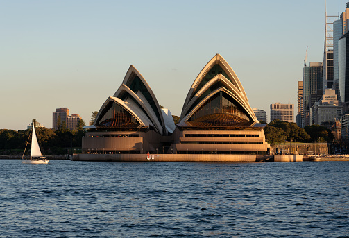 Sydney Australia - August 2, 2019: A sunny day in Sydney, Australia, and a small sailing boat crosses the blue waters of Sydney Harbour. Beyond it can be seen the iconic shape of the Opera House.