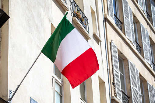 Close-up view of an Italian flag placed on a window sill with red and white flowers.The facade of the old building is painted ochre-coloured. Square composition and copy space.