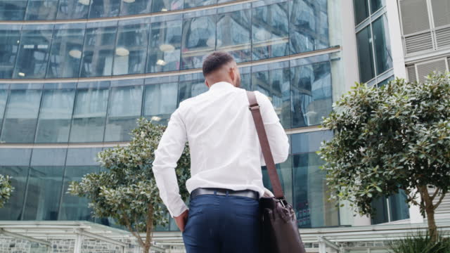 Rear view of young businessman walking alone in a city. Corporate professional carrying bag while commuting to or from work. Low angle of male executive looking around exploring business district