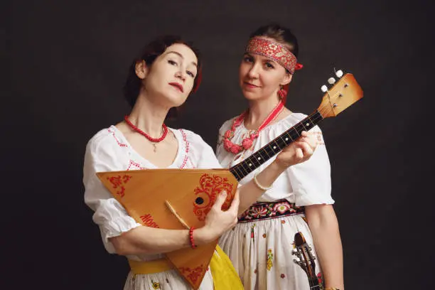 Women musicians in Russian folk dresses with musical instruments on a black studio background. Happy artists from Russia in white national clothes with stringed musical instruments