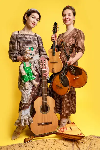 Photo of Women musicians in dresses with musical instruments on a yellow studio background. Happy artists with stringed musical instruments with smiles on faces