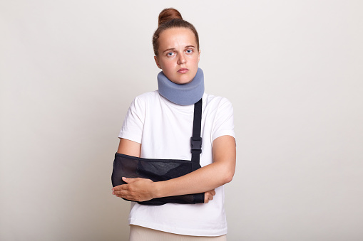 Image of sick injured woman with bun hairstyle wearing casual T-shirt and Shants collar, looking at camera, holding her hands, felling pain, standing in arm sling isolated on light gray background.