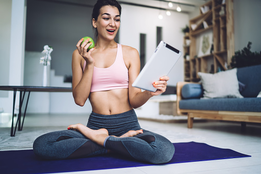 Full body of smiling brunette in pink sports bra and grey leggings browsing tablet and looking at screen happily while sitting in lotus pose with green apple on yoga mat