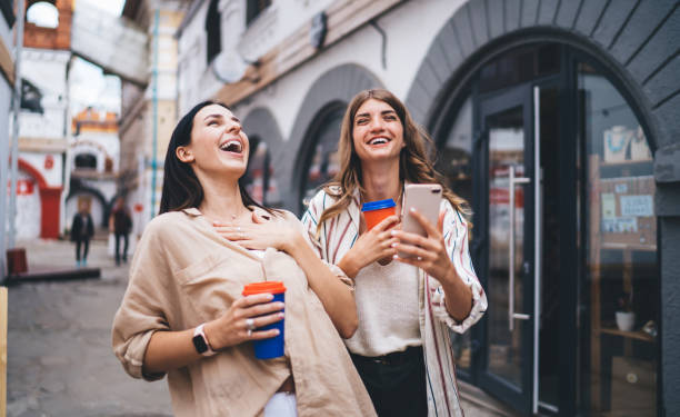 Excited female friends smiling broadly especially in unrestrained manner during cellphone networking, grinning hipster girls with smartphone and coffee to go satisfied with meeting for mobility amigas emocionadas sonriendo ampliamente, especialmente de manera desenfrenada durante las redes de teléfonos celulares, sonriendo a las chicas hipster con teléfono inteligente y café para ir satisfechas con la reunión para la movilidad.