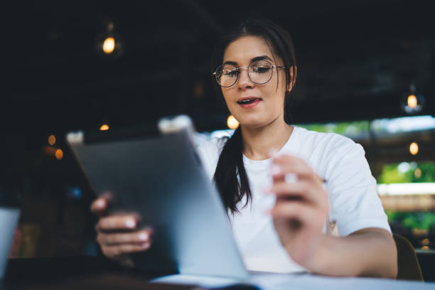 istock Skilled caucasian female in eyewear for vision correction installing new application on digital tablet for learning,smiling woman 20s blogger share media and multimedia content in blog sitting in cafe 1407266062