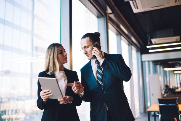 Caucasian boss reading financial news on modern touch pad while communicating with corporate investor via cellphone, formally dressed colleagues using technology with 4g internet in office interior