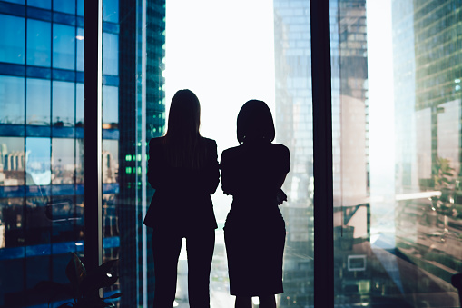 istock Back view of female colleagues in formal wear standing near window looking at modern exterior of skyscrapers in business center, silhouette of women together planning future success of brainstorming 1407268206