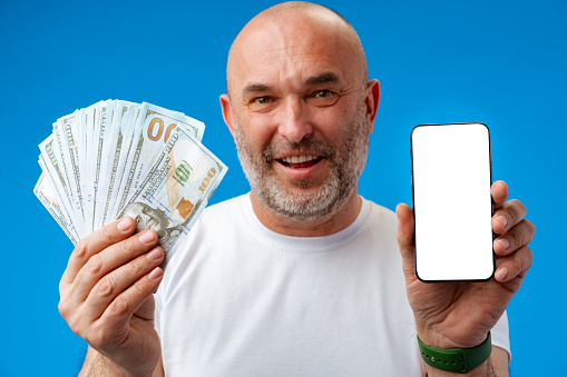 Handsome smiling man holding dollar banknotes and smart phone with white blank screen in studio