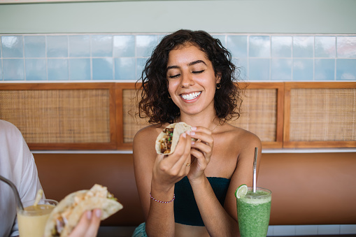 Positive young Hispanic lady in summer outfit smiling while eating tasty Mexican beef taco and drinking lime smoothie in cozy cafe