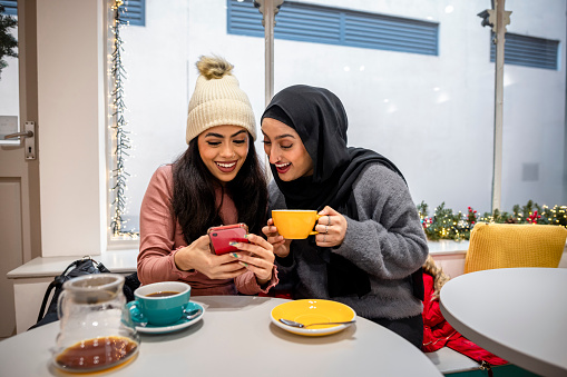 A medium shot of two young women wearing warm casual clothing and accessories on a winter's day in Newcastle Upon Tyne. They are sitting at a table in a coffee shop that is decorated with Christmas decorations, chatting and laughing while using a smartphone.