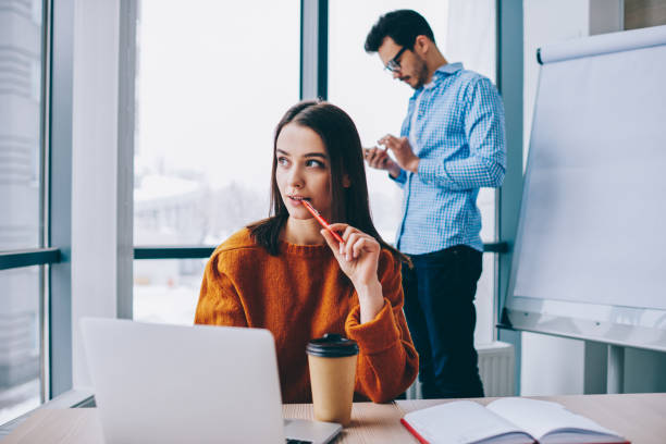 istock Selective focus on skilled female employee sitting at table desktop and thinking about laptop projecting while blurred male colleague on background using smartphone for banking, office workspace 1407272326