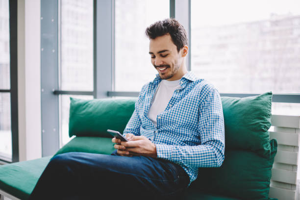 istock Cheerful hipster guy dressed in casual wear writing email message via application on cellular phone resting on comfortable couch, successful male blogger searching information at online platform 1407272366