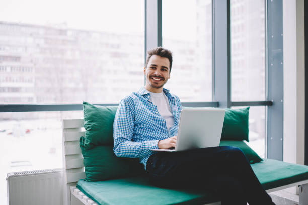 istock Cheerful male freelancer with modern laptop device sitting at comfortable couch and smiling, happy Caucasian IT professional in smart casual wear waiting for netbook update for working in online 1407272370