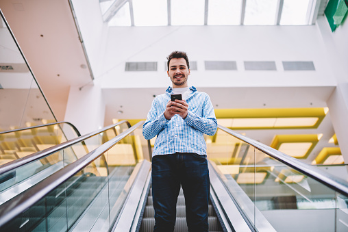 Half length portrait of cheerful Caucasian man smiling at camera while going down on modern escalator holding smartphone for checking balance on credit card via application before shopping