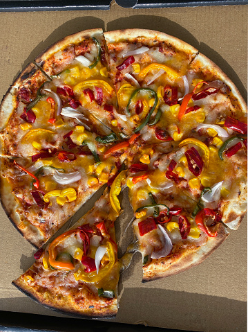 Stock photo showing close-up, elevated view of cardboard takeaway pizza box containing a sliced red, yellow and green pepper, red onion and sweetcorn pizza, topped with a rich tomato sauce and melted buffalo mozzarella.