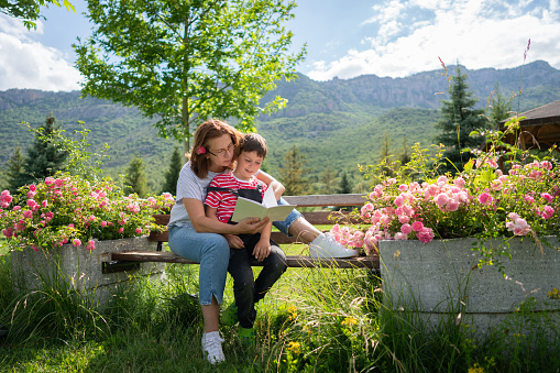 Mother and son are reading a book outdoor on the bench with rose flower and mountain background