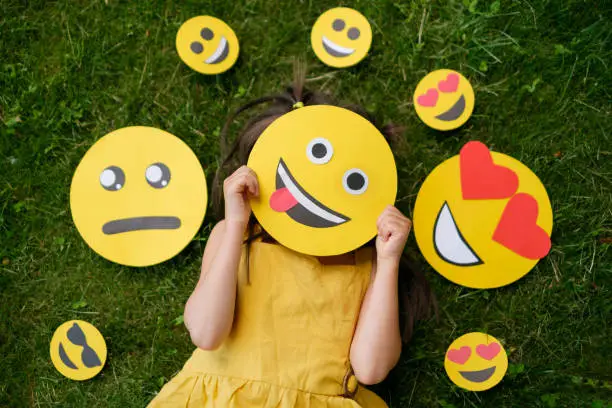 The child covers his face with a emoticon showing his tongue. The person is lying on the grass with emoticons with different psychological moods.