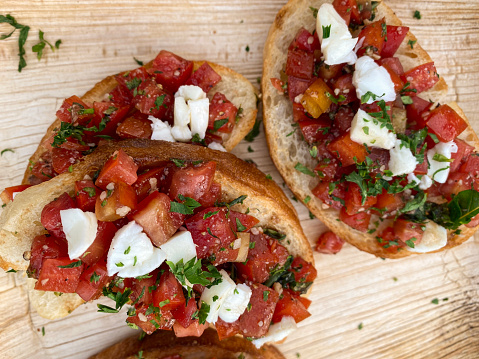 Stock photo of bruschetta recipe open sandwich with baguette bread slices, garlic and basic pesto sauce on diced chopped cherry tomatoes, sliced red onion, mozzarella cheese, and fresh basil leaves on wood grain background.