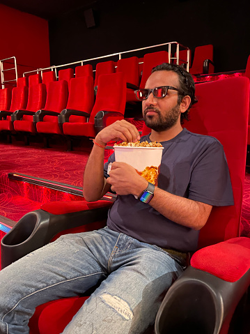 Stock photo showing close-up view of man sitting in aisle seat of cinema, wearing 3D glasses to watch a film whilst eating popcorn from a tub.