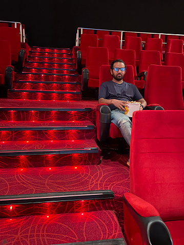 Stock photo showing close-up view of man sitting in aisle seat of cinema, wearing 3D glasses to watch a film whilst eating popcorn from a tub.