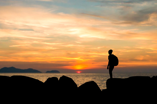 Silhouette of young man with backpack standing on rock looking beautiful view sunset over the sea at sunset.