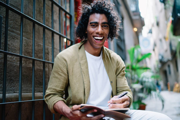 istock Half length portrait of cheerful hipster guy 20s enjoying working outdoors making notes for article publication, smiling young male designer creating sketches looking at camera sitting on city street 1407276967