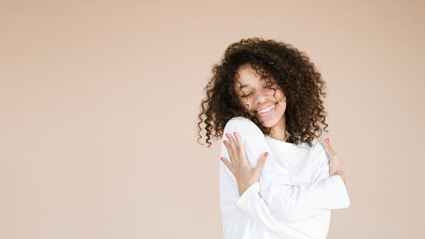 African american girl holding hands on shoulders Photo of smiling african american girl holding hands on shoulders wear casual white t-shirt isolated beige background hugging self stock pictures, royalty-free photos & images