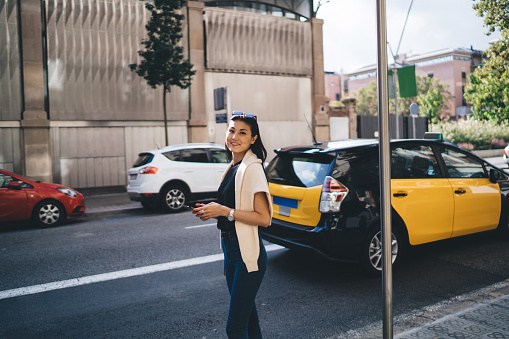 Half length portrait of beautiful woman traveler in casual wear standing on road using mobile phone for booking taxi, asian hipster girl looking at camera waiting for cab transporting in megalopolis