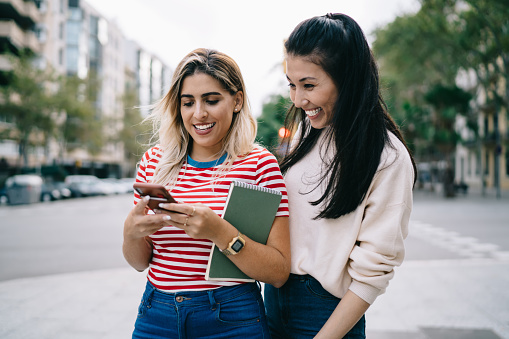 Joyful hipster girls reading funny publication during social networking via modern cellular technology enjoying together blogging in city, smiling female tourists tracking mobile gps during travelling
