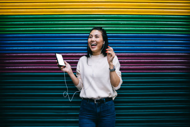 Excited Asian user with blank smartphone grinning at publicity area satisfied with free time for listening funny audio podcast, joyful woman in earphones laughing and rejoicing at coppy space area usuario asiático emocionado con teléfono inteligente en blanco sonriendo en el área de publicidad satisfecho con el tiempo libre para escuchar podcasts de audio divertidos, mujer alegre con auriculares riendo y regocijándose en el área del espacio cop