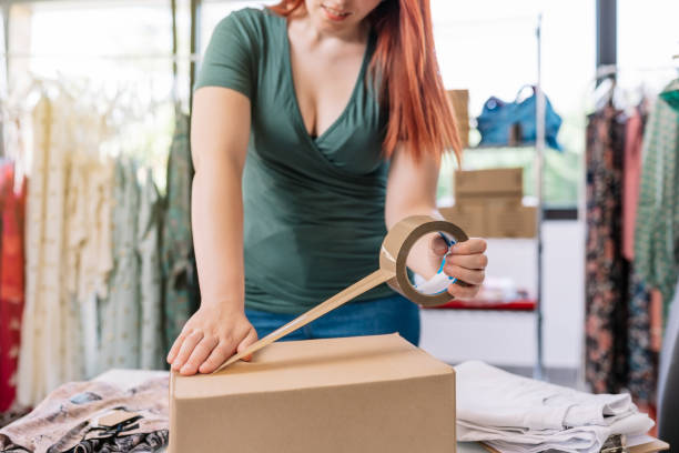 cutaway view of a young businesswoman, packing a box with tape, for a shipment from her online clothing shop. young woman preparing a package for a client in the office. work and business concept. - sales clerk store manual worker retail imagens e fotografias de stock