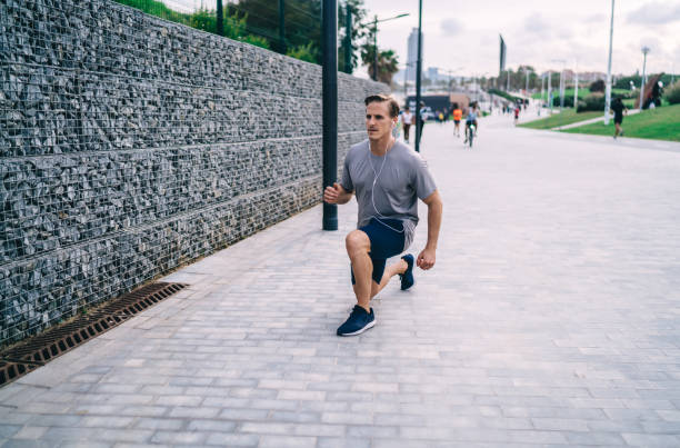 istock Caucasian athletic bodybuilder in electronic headphones squatting during morning workout training at street, sportsman 30 years old in earphones listening music podcast while stretching legs muscles 1407281790