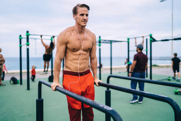 Positive caucasian strong athlete doing exercises on bars in gym outdoors enjoying listening music keeping healthy lifestyle,male bodybuilder training muscles and body shape topless in earphones atleta fuerte caucásico positivo haciendo ejercicios en barras en el gimnasio al aire libre disfrutando de escuchar música manteniendo un estilo de vida saludable, culturista masculino entrenando músculos y forma corporal en topless en auriculares