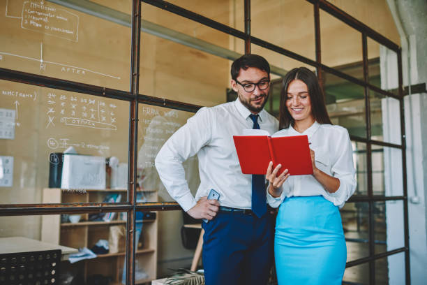Successful male and female professionals 30s reading information checking from notepad, happy confident marketing employees analyzing report noted in paper textbook standing in office interior