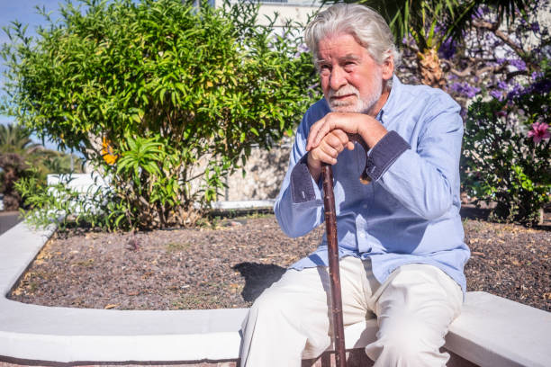 istock Old senior man with hands over the cane sitting in public park with sad expression. Elderly retiree with back pain using walking stick. Tree and flowers on background 1407283563