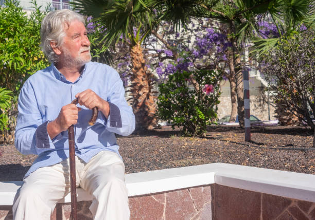 istock Old senior man with hands over the cane sitting in public park with sad expression. Elderly retiree with back pain using walking stick. Tree and flowers on background 1407283627
