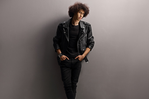 Young man with a leather jacket and curly hair leaning on a gray wall and looking at camera