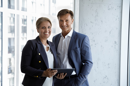 Middle-aged businesspeople in formal elegant suits pose in office with digital tablet smile staring at camera look satisfied. Professional staff members, executive male female manager portrait concept