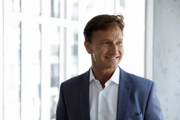 Head shot of successful middle-aged handsome businessman in elegant formal suit smile looks aside pose at modern skyscraper office. Executive manager, company owner or project leader portrait concept
