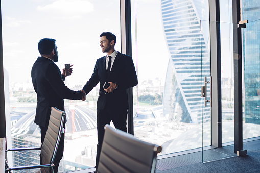 Well dressed businessmen getting acquainted with each other while standing in front of panoramic window in modern office space and shaking hands