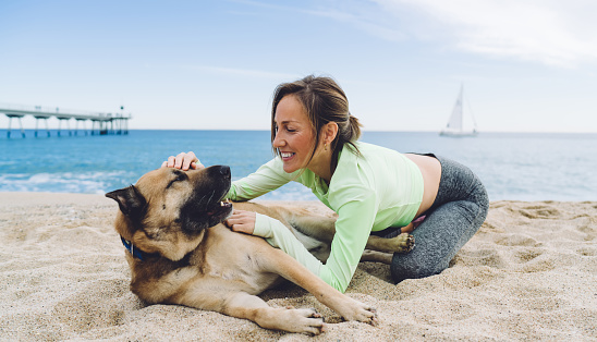 Joyful fit woman having fun with adorable mongrel dog with dark muzzle while spending leisure time at coastline seashore, playful female runner in active tracksuit stroking cute doggie pet
