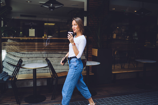 Beautiful woman in white top and blue jeans with notebook in hand walking on street and drinking coffee against backdrop of city cafe